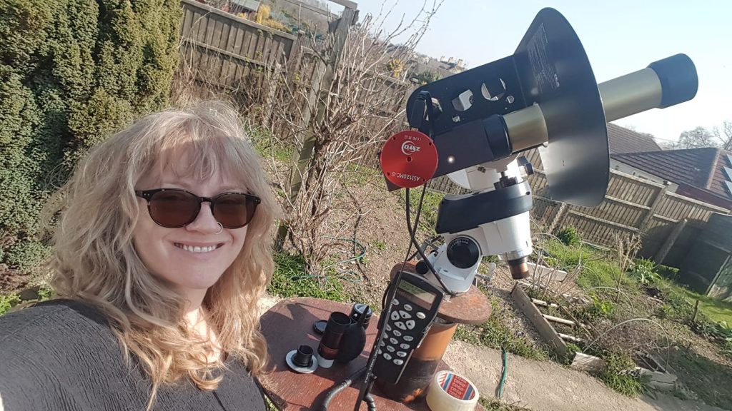 Mary standing next to a telescope pier in her garden. A Coronado PST solar telescope is on the mount and an ASI120MC camera is attached to the telescope